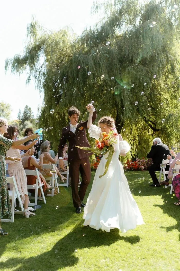 A bride and groom dancing down a grass aisle under a willow tree as guests blow bubbles